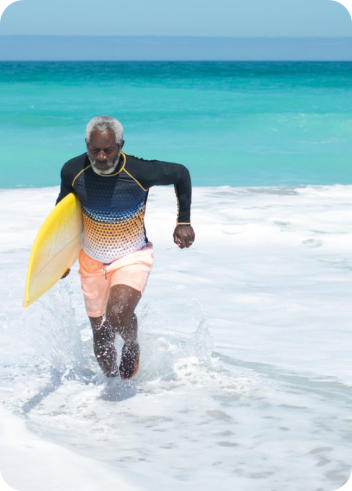 A senior man is running out of the waves onto the beach carrying a surfboard under his arm.