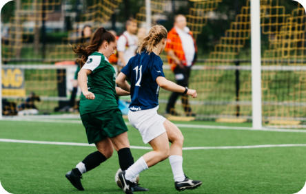 Two teenage girls wearing different soccer uniforms are on the soccer pitch both trying to kick the ball.